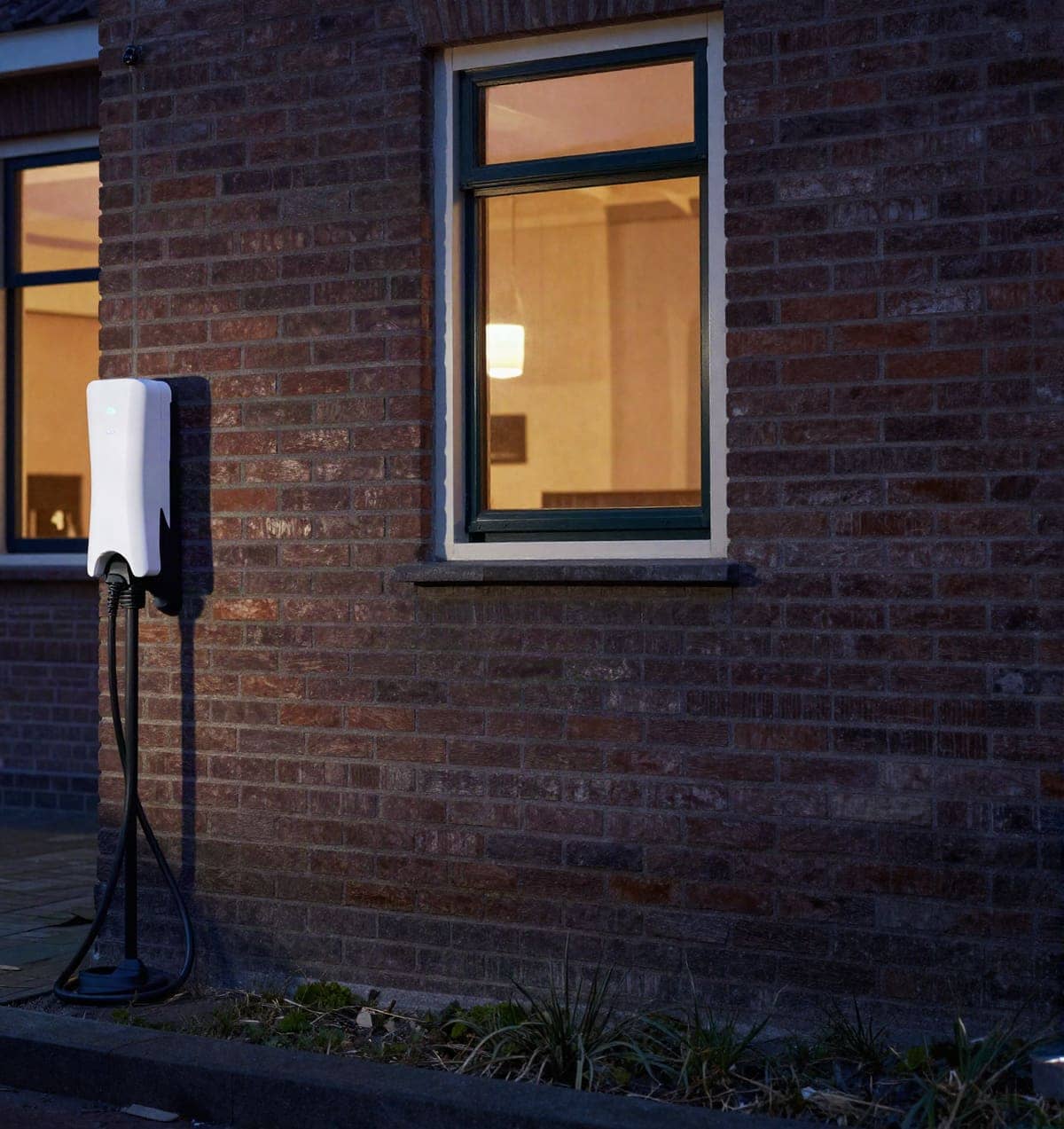 Laadpaal aan een woning in de avond, met licht achter de ramen